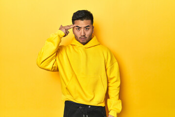 Fototapeta premium Bold Asian man in yellow hoodie on yellow studio pointing temple with finger, thinking, focused on a task.