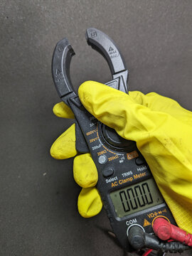 Hand in yellow glove holding a clamp meter showing zero reading on display