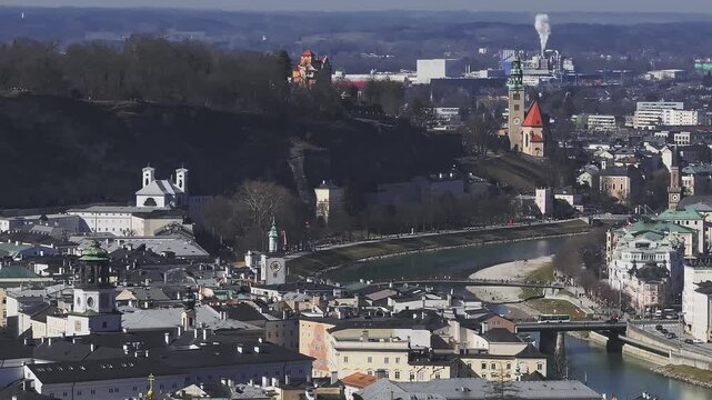 Aerial view of Salzburg, Austria, showing the Salzach River, St. Andra Church, historic buildings, moving vehicles, and distant industrial smoke.