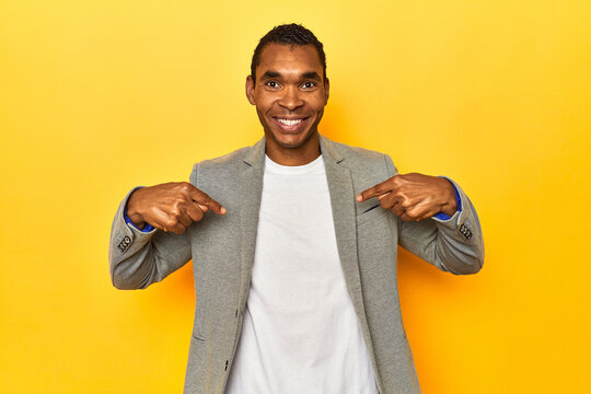 African American man in casual blazer, yellow studio, surprised pointing with finger, smiling broadly.