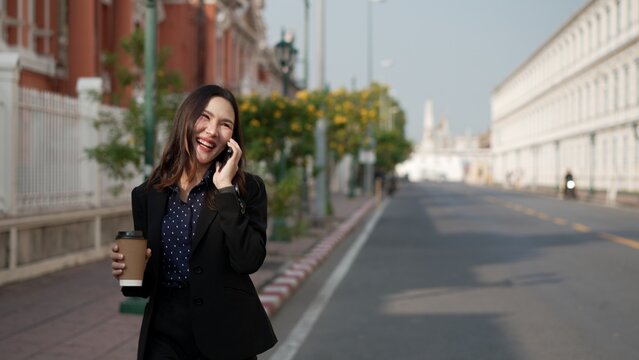 asian businesswoman walking on street laughing while talking on mobile phone and holding coffee cup