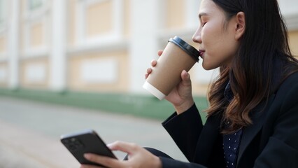closeup side view of young asian woman drinking coffee and holding smartphone outdoors