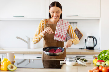 Young lady housewife cooking meat, frying on pan and salting it, woman wearing apron while...