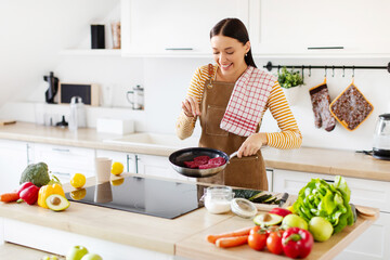 Pretty young lady frying meat and seasoning with salt, cooking dinner in kitchen interior, trying new recipe and smiling