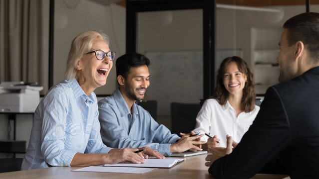 Happy lady leader laughing at diverse corporate board meeting. Business people communicate, executive older lady manager talking with colleagues, enjoy office life. International team having fun