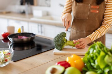 Close-up of woman in apron cutting broccoli on chopping board, preparing ingredients for healthy...