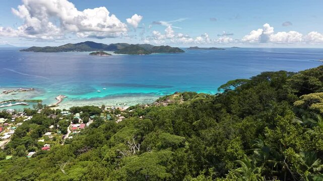 La Digue, Seychelles, viewed from above with a vibrant coastal village, turquoise waters, lush forest, and distant mountainous islands under a bright blue sky.
