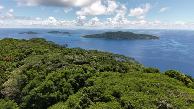 La Digue, Seychelles, viewed from above with a vibrant coastal village, turquoise waters, lush forest, and distant mountainous islands under a bright blue sky.
