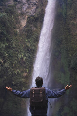 Back view of a man in blue hoodie raising his arms in front of a waterfall. Freedom traveling.