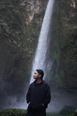 Asian man in black hoodie confidently standing in front of a waterfall