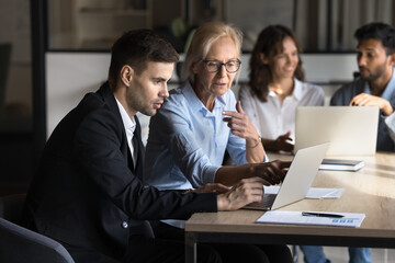 Two diverse corporate professionals teamwork with laptop on sales or technological project. Experienced executive mid-aged businesswoman mentors young coworker utilizing computer in modern workplace