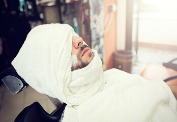 Mature male with beard receiving professional grooming, reclining in barbershop chair with hot towel covering face and shaving cream applied, preparing for precise trimming