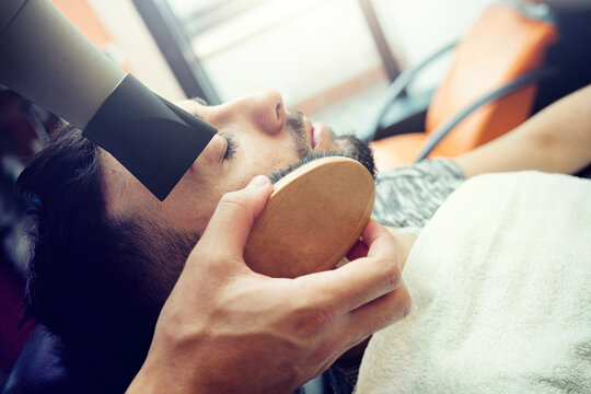 Professional barber styling client's beard with hairdryer and brush, working at grooming station in traditional barbershop interior