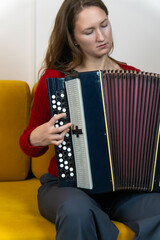 Young musician sitting on a yellow sofa, playing a bayan accordion and practicing her musical...