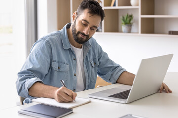 Remote work lifestyle. Confident millennial businessman entrepreneur freelancer engaged in business planning take brief notes write down information ideas from notebook screen sitting at office desk