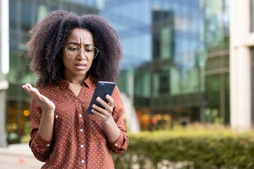 A frustrated woman in glasses looks at her smartphone, seemingly upset with the content she is...