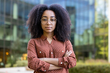 A confident woman with curly hair stands in front of a modern building, arms crossed, looking...