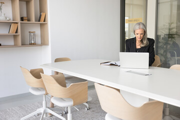 Businesswoman at work. Focused older woman ceo corporate executive manager wear formal suit engaged in paperwork use notebook to prepare for conference at table in empty office boardroom. Copy space