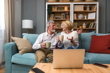 Senior couple sitting at home on sofa, watching news, movie or sport game on laptop computer. Mature married people, man and woman, using technology for entertainment. Simple living concept.