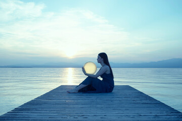 elegant woman in backlight embracing a moon-shaped lamp in front of a marine sunset