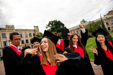 Happy group of young graduates celebrating in academic caps and gowns outdoors on a sunny day,...