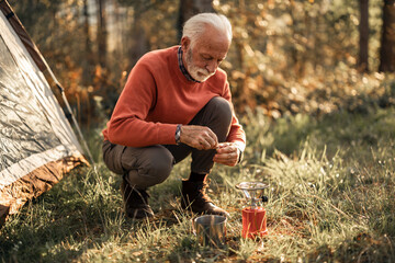 Senior Man Camping Outdoors Preparing a Portable Stove Near a Tent in Forest