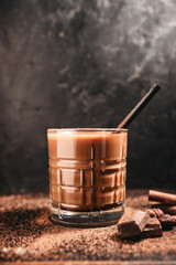 Hot chocolate, milkshake, cocoa drink in a glass with a straw, almonds, hazelnuts, cinnamon and chocolate pieces on a dark wooden background