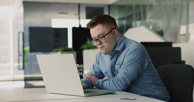 Serious down syndrome manager man in glasses working on project in company with inclusion, diversity, typing on laptop at workplace, staring at screen, thinking on solution, speaking