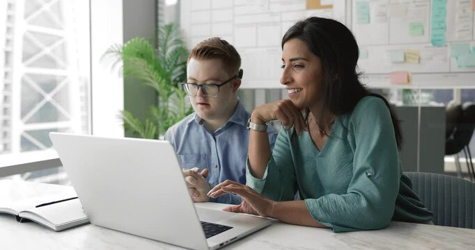 Serious young down syndrome professional man and positive Latin businesswoman working at laptop together, discussing online product presentation, inclusive company corporate app