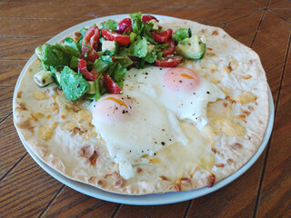 Breakfast plate featuring poached eggs on a flatbread with fresh salad served on a rustic wooden table