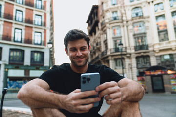 Smiling young man browsing phone in urban square, dressed in black, relaxed after morning workout, enjoying digital entertainment or online shopping on mobile device