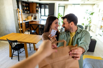 Young smiling couple receiving grocery bag from delivery person and paying with credit card, food delivery service concept