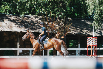 A young equestrian rides a horse in an outdoor stable, enjoying a sunny day. The scene captures the...