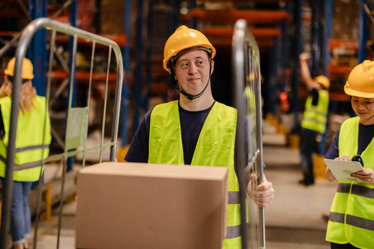 Young warehouse worker with down syndrome pushing trolley with cardboard box