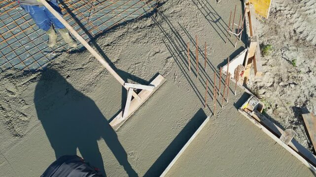 Construction workers leveling fresh concrete floor with a wooden tool at construction site. Pouring concrete into a formwork with reinforcement and leveling for building a new house, slow motion