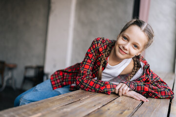 Pretty 10 years old girl with braided hair in red plaid shirt resting against wooden windowsill by...