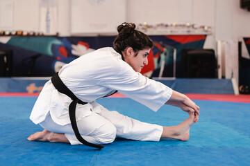 woman in a white martial arts uniform with a black belt sits on a blue mat and stretches forward to touch her foot inside a brightly lit training facility
