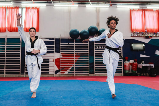A young woman and man wearing white taekwondo uniforms with black belts perform synchronized martial arts movements on a blue mat inside a brightly lit training hall