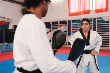 A female martial artist in uniform and black belt stands ready in front of a male partner holding training pads inside a gym with red curtains and mat flooring