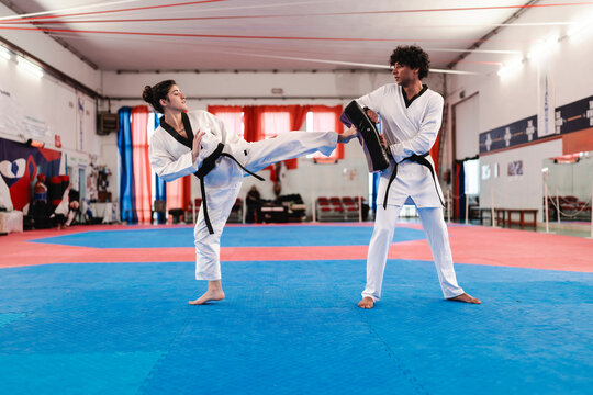 A female martial artist in white uniform and black belt executes a controlled side kick toward a training pad held by a male partner inside a red and blue mat dojo