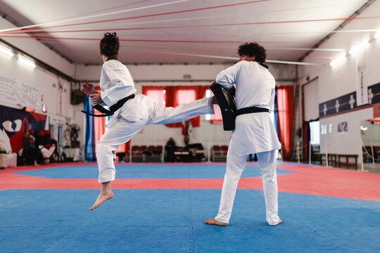 A woman in a martial arts uniform and black belt leaps off the ground to strike a held training pad with her foot during a dynamic taekwondo practice session indoors