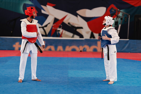 A male and female martial artist wearing full protective gear including chest guards and helmets stand facing each other on a blue mat inside a brightly lit gym