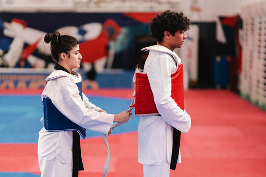 A female martial artist wearing a blue chest protector helps tie the red chest guard of her male partner in a training gym with red and blue mats and background mural
