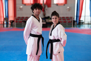 A male and female martial artist in white uniforms and black belts stand side by side on a blue mat in a taekwondo gym with red curtains and blurred seating in the background