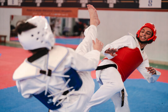 A female martial artist in blue protective gear prepares to block a high kick from her male opponent during a sparring session on a red and blue mat inside a gym