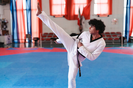 A male martial artist in a white uniform and black belt performs a high side kick on a blue mat inside a taekwondo gym with red curtains and chairs in the background
