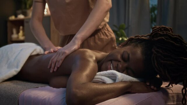 African American woman smiling, while unrecognizable masseuse gently rubbing her back during massage session in spa