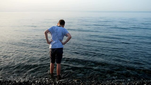 Teen boy standing in sea on pebble beach looking at transparent clear water. Teenger on vacation admiring seascape at sunset. Travel, tourism, wanderlust. Teenage guy resting relaxing on sea coast.
