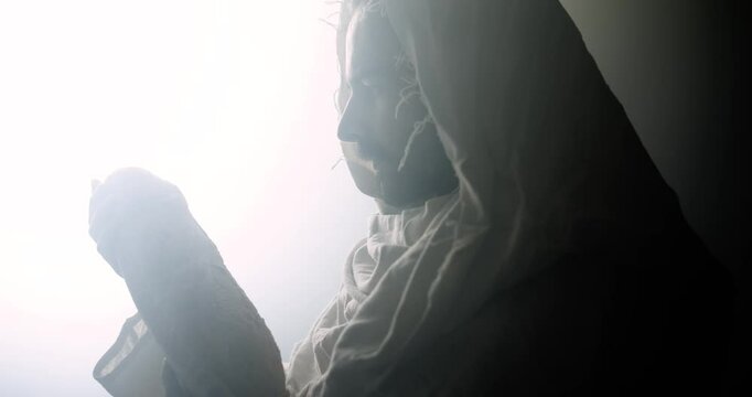 Closeup of Jesus Christ in white hooded robe holding bread as he stands in glowing, bright light. Perfect for representation of Jesus as the bread of life, last supper, communion, the gospel.
