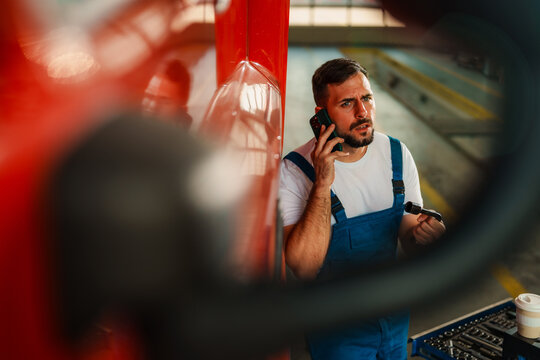 Mechanic on a break leans against a red truck, talking on the phone while holding a coffee cup. Tools and a toolbox are visible in the background, suggesting a workshop environment.